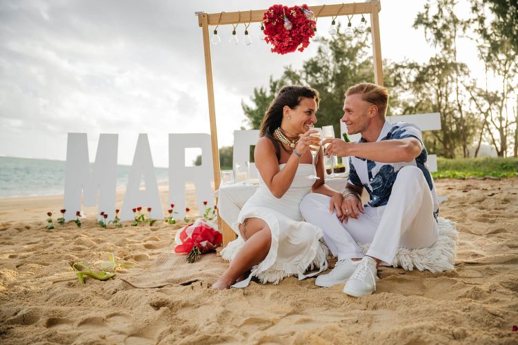 couple sat on beach with champaign, marry me sign behind them