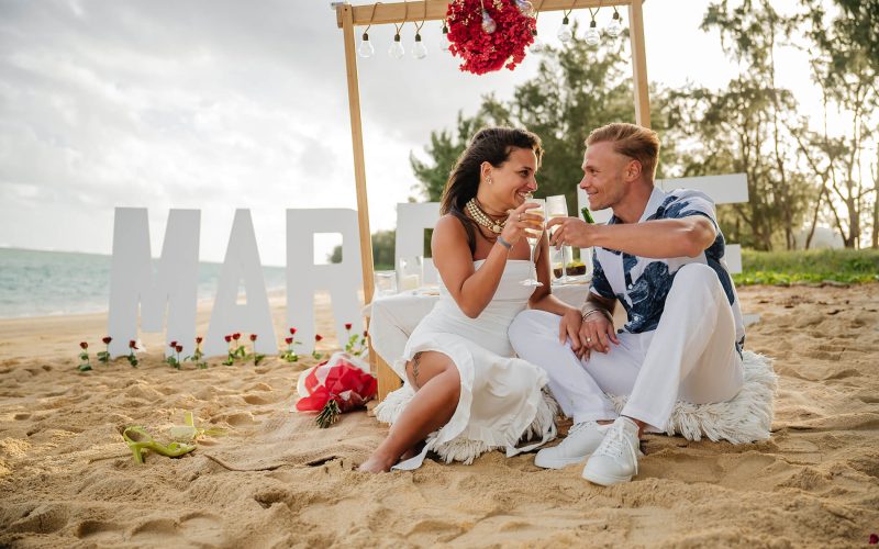 couple sat on beach with champaign, marry me sign behind them